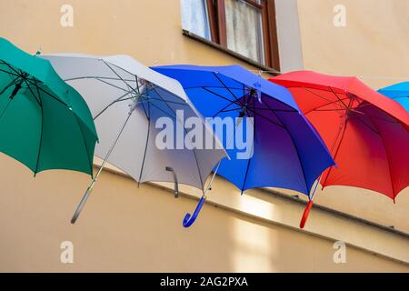 Umbrellas in different colors against the backdrop of a wall on a sunny summer day. Green, gray, blue and red umbrellas hang in the air against the ye Stock Photo