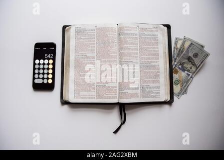 Overhead shot of an open bible in the middle of money and a smartphone on a white surface Stock Photo