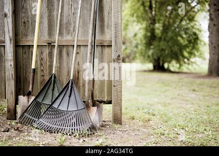 Closeup shot of two leaf rakes and shovels leaned against a wooden fence with a blurred background Stock Photo