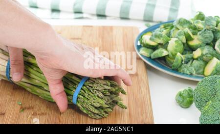 Asparagus, broccoli and Brussels sprouts on a wooden cutting board with white background, close up, woman hands Stock Photo
