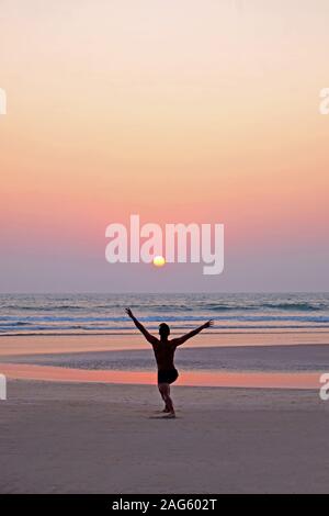 An unrecognizable man doing a yoga pose, the sun salutation on a wide open empty sandy beach, he is kneeling with his arms outstretched, he is lined u Stock Photo