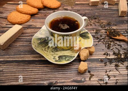 A Cup of black tea, tea leaves, pieces of brown sugar, oatmeal cookies, waffles on a wooden background. Close up Stock Photo