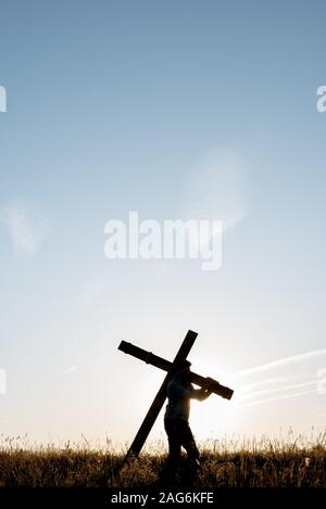 Vertical shot of a male carrying a handmade wooden cross in a grassy field under a blue sky Stock Photo