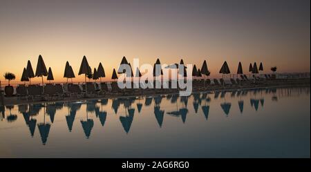 Lot of closed parasols and folding chairs on the beach reflected in the sea during sunset Stock Photo