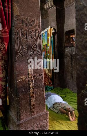 Ethiopia, Amhara Region, Lalibela, inside Bet Maryam Church, devotee prostrating in prayer beyond stone pillar with cruciform carved painted design Stock Photo