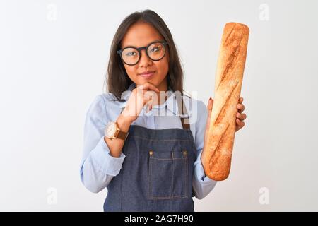 Young chinese baker woman wearing glasses holding bread over isolated white background serious face thinking about question, very confused idea Stock Photo