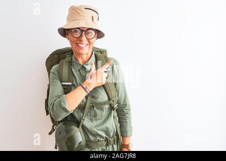 Middle age hiker woman wearing backpack canteen hat glasses over isolated white background cheerful with a smile of face pointing with hand and finger Stock Photo