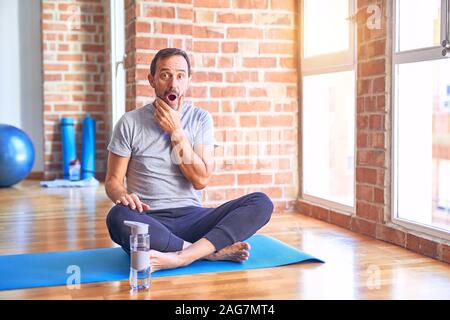 Middle age handsome sportman sitting on mat doing stretching yoga exercise at gym Looking fascinated with disbelief, surprise and amazed expression wi Stock Photo