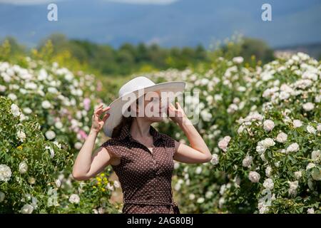 Beautiful woman in retro style polka dot dress and hat staying in rose field during spring warm sunny day with blue cloudy sky. The concept of Bulgari Stock Photo