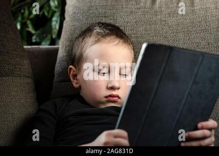 Young boy sitting on sofa in living room and watching cartoons on tablet. Portrait of a smart pre-school child using devices at home. Modern kid.