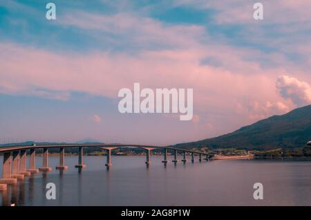 Beautiful scenery of a concrete bridge over the lake near high mountains during sunset in Norway Stock Photo