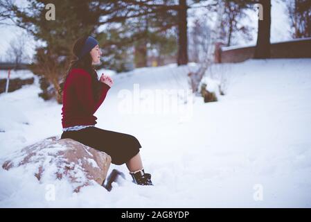 Dark-haired female with a red jacket and a skirt sitting on a stone and praying in a snowy area Stock Photo