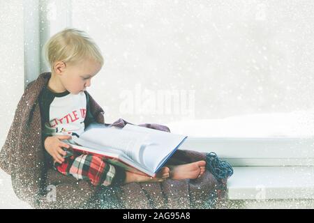 Child reads book by the window. Beautiful winter weather with snow falling Stock Photo