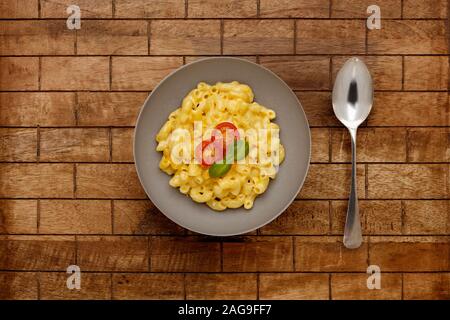 A bowl of delicious Macaroni and cheese, topped with cherry tomatoes and basil, on a checked wooden background, with a spoon. Stock Photo