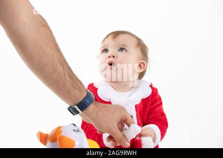 Stock studio photo with white background of a baby disguised as Santa Claus sitting on the floor while an adult hand grabs his hat from his hands. Stock Photo