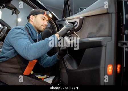Car service worker in uniform provides a professional vehicle interior cleaning, wiping door panel with a brush at the car service station