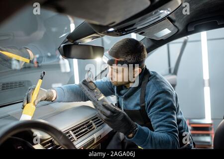 Car service worker in uniform provides a professional vehicle interior cleaning, wiping front panel with a brush at the car service station