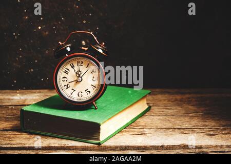 Old retro alarm clock on green book in rustic interior with flying dust. Stock Photo