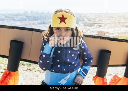 portrait of a funny little child girl disguised as a superhero scolding the bad guys with homemade costume and cardboard plane wings in front of a cit Stock Photo