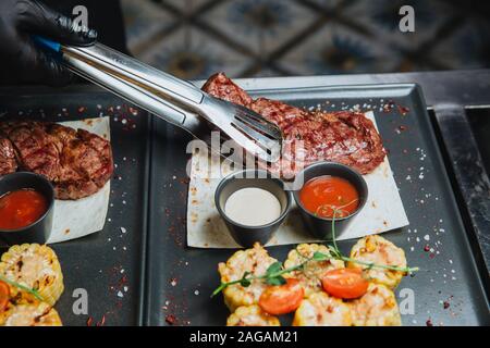 The cook puts tongs in a plate of juicy fried steak with two sauces . Hot Meat Dishes. Stock Photo
