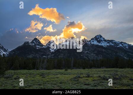 Clouds colored by orange sunset behind the peks of the Grand Teton mountain range with contrasting dramatic light and deep shadows Stock Photo