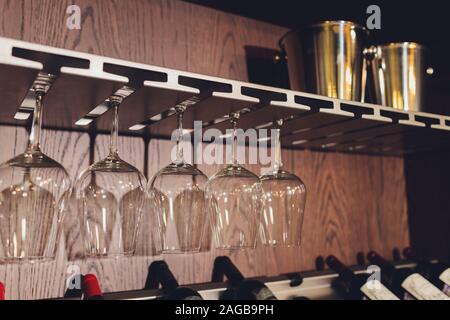 Closeup many upside down empty clear transparent crystal upturned wine glasses hanging in straight rows on brown wooden shelf, rack, showcase, stainle Stock Photo