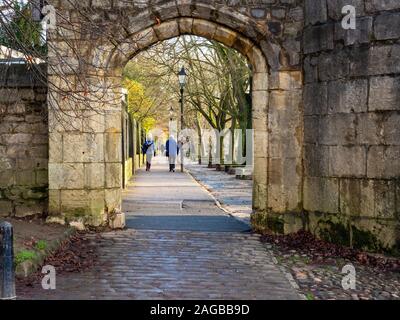 View through a stone arch to a couple strolling on the tree lined riverside Dame Judi Dench Walk in York, England Stock Photo