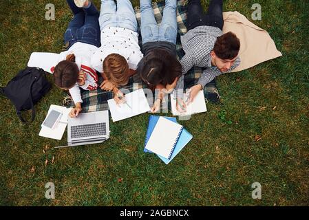 Writing on paper. With laptop. Group of young students in casual clothes on green grass at daytime Stock Photo