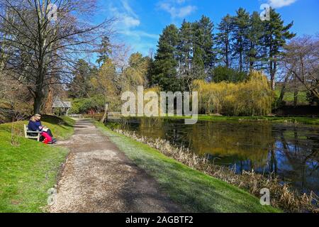 A couple sitting on a wooden bench having a picnic with the wooden Boat House in the background at Bodnant Gardens, Tal-y-Cafn, Conwy, Wales, UK Stock Photo