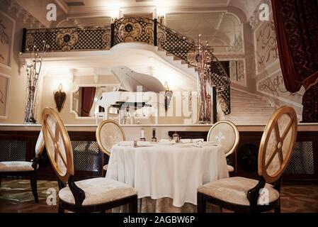 Table for the four people is ready to receive rich visitors. Interior of luxury restaurant in vintage aristocratic style with piano on the stage Stock Photo