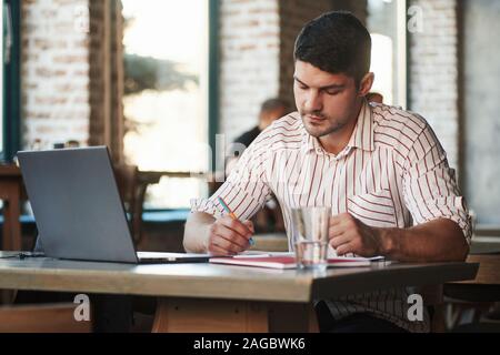 Guy making some notes in his textbook. Adult man sits in cafe at daytime and using the laptop for the remote work Stock Photo
