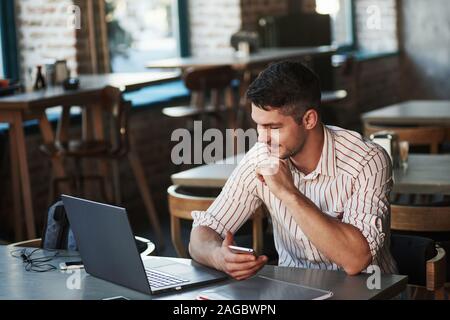 Looks like everything is going nice. Adult man sits in cafe at daytime and using the laptop for the remote work Stock Photo