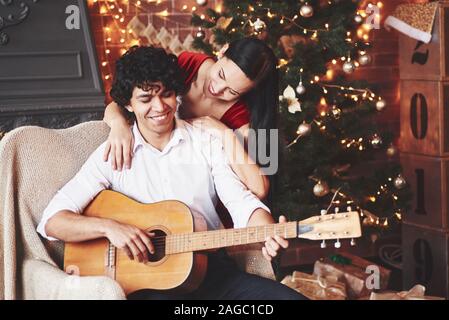 Holds acoustic guitar. Beautiful couple celebrating New year in the decorated room with Christmas tree behind Stock Photo