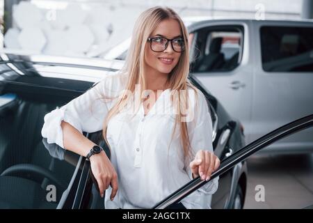 With van behind. Cute girl in eyewear stands near the car in auto saloon. Probably her next purchase Stock Photo