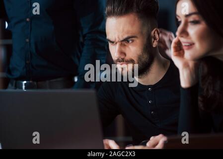 Focused photo on the unshaved guy. Team of young business people works on their project at night time in the office Stock Photo