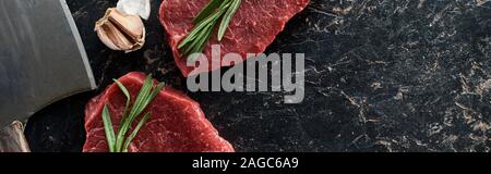panoramic shot of unprocessed beef steaks with rosemary sprigs near garlic and knife on black marble surface Stock Photo