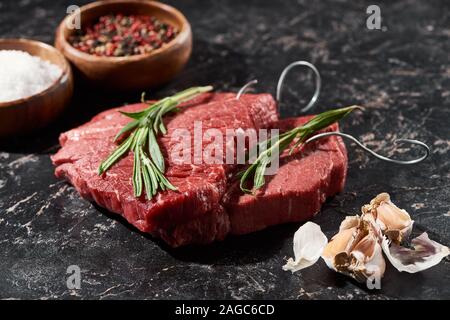 raw beef steak with rosemary near garlic cloves and small bowls with salt and peppercorns on black marble surface Stock Photo