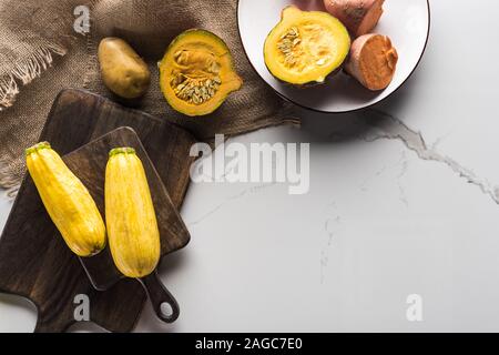 top view of wooden cutting boards with zucchini, potato, pumpkin and plate with yam on marble surface with hessian Stock Photo