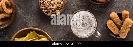 panoramic shot of mug of light beer near peanuts and crisps on brown textured surface Stock Photo