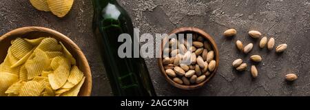 panoramic shot of bottle of beer near bowls with pistachios and crisps on brown textured surface Stock Photo