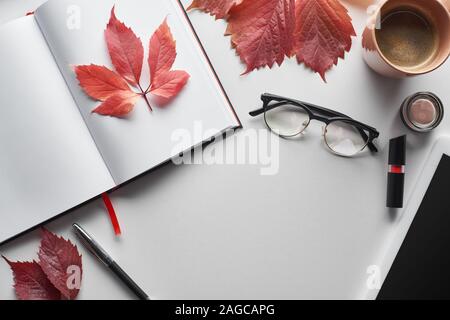 top view of notebook near glasses, coffee cup, cosmetics, pen and red leaves of wild grapes on white table Stock Photo