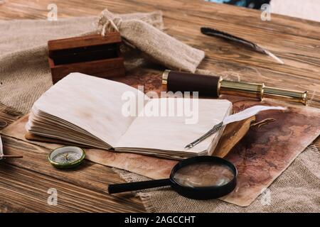 open copy book with blank pages near telescope, compass and magnifying glass on wooden table with hessian and world map Stock Photo