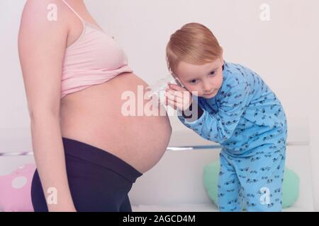 Listening to tummy. Curious handsome dark-eyed boy feeling very interested while listening to pregnant tummy of his pregnant mother. Stock Photo