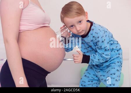 Listening to tummy. Curious handsome dark-eyed boy feeling very interested while listening to pregnant tummy of his pregnant mother. Stock Photo