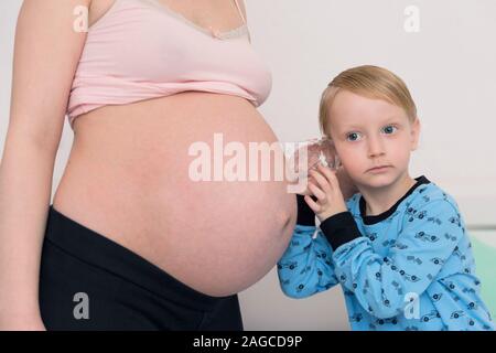 Listening to tummy. Curious handsome dark-eyed boy feeling very interested while listening to pregnant tummy of his pregnant mother. Stock Photo