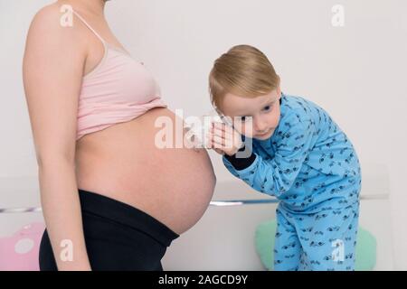 Listening to tummy. Curious handsome dark-eyed boy feeling very interested while listening to pregnant tummy of his pregnant mother. Stock Photo