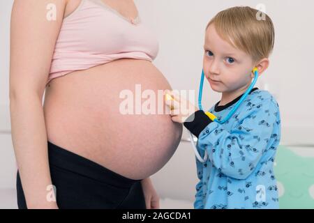 Listening to tummy. Curious handsome dark-eyed boy feeling very interested while listening to pregnant tummy of his pregnant mother. Stock Photo