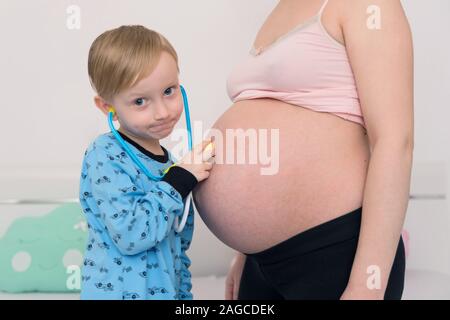 Listening to tummy. Curious handsome dark-eyed boy feeling very interested while listening to pregnant tummy of his pregnant mother. Stock Photo
