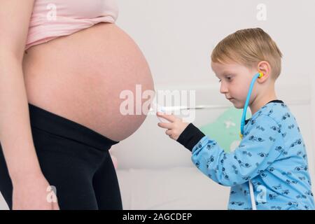 Listening to tummy. Curious handsome dark-eyed boy feeling very interested while listening to pregnant tummy of his pregnant mother. Stock Photo