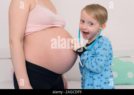 Listening to tummy. Curious handsome dark-eyed boy feeling very interested while listening to pregnant tummy of his pregnant mother. Stock Photo